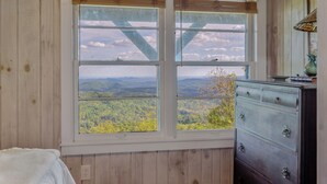 Ein Zimmer mit einem großen Fenster, das einen Blick auf eine bewaldete Landschaft bietet. Es gibt eine Kommode mit Schubladen und eine Lampe darauf.