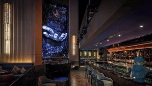 A bar area with a large digital screen displaying a blue abstract image, bar stools, and a variety of bottles on shelves. - Burj View Studio Downtown (Dubai)