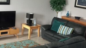 A living room with a black leather sofa, a wooden side table with a lamp, a TV, and a small plant. - Sea Surf, 2 bedroom, fully equipped ground floor apartment, sleeps 4, in city (Bangor)