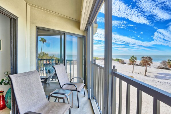 A balcony with a view of the beach, two chairs, and a table.