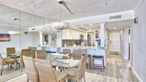 A modern kitchen with white cabinets, a central island, and a dining area with wicker chairs and a glass table. - 🏖️Beachfront🌴Heated Pool, Hot Tub-Direct Beach Access-Land's End 10-303🌊 (Treasure Island)