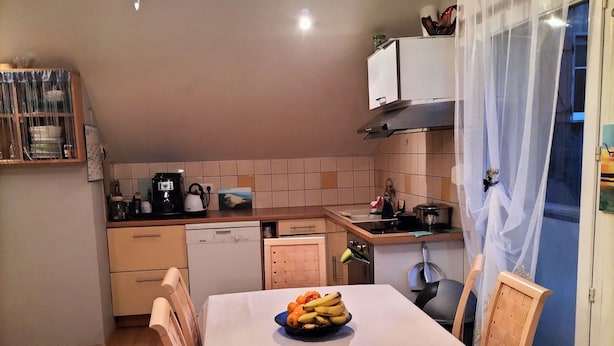 Un coin repas avec une table dressée pour deux personnes, un lustre et une cuisine en arrière-plan.