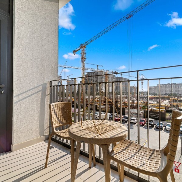 A balcony with two wicker chairs and a small round table, overlooking a construction site with cranes and buildings.