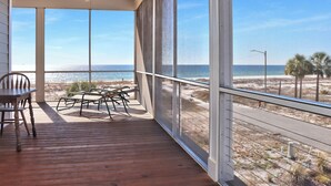 A balcony with a wooden floor, a table, and chairs, overlooking a beach and ocean. - 🌊Beach View-2 Beachfront Screened Decks-4BR-Windchimes🌊 (Mexico Beach)