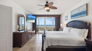 A bedroom with a bed, a ceiling fan, a TV, a dresser, and a view of the ocean.