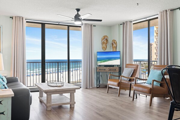 A spacious living room with a large window overlooking the beach, a ceiling fan, and a flat-screen TV.
