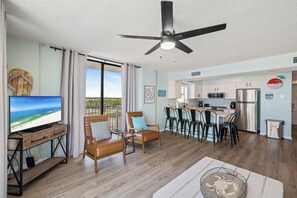 A modern living room with a TV, seating, a ceiling fan, and a view of the beach.