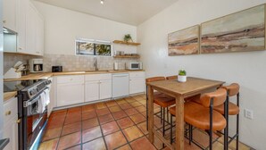 Une cuisine avec des meubles blancs, un plan de travail en bois, un sol carrelé, une table à manger avec des chaises et un tableau accroché au mur.
