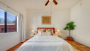 Une chambre à coucher avec un grand lit, deux tables de chevet avec des lampes, un ventilateur de plafond, une fenêtre avec des rideaux, une plante en pot et un élément décoratif mural.