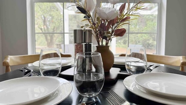 Una mesa de comedor dispuesta con platos, vasos y cubiertos, con un florero y una botella de agua encima.