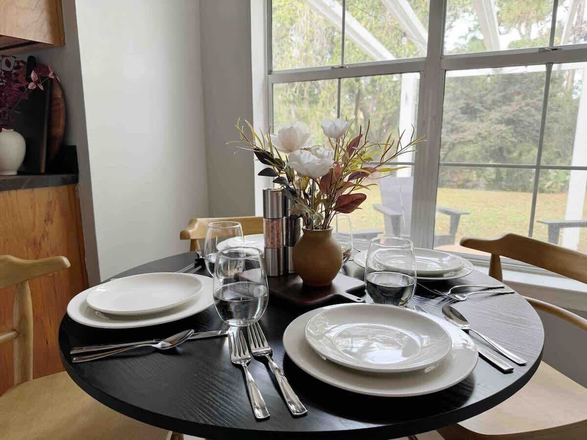 A dining table set for four with white plates, cutlery, and glasses, a vase with flowers, and a view of greenery through a window.