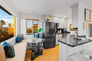 A modern kitchen with a large refrigerator, a dining area with a glass table, and a view of palm trees outside.