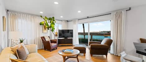 A modern living room with a sofa, armchairs, a coffee table, and a TV. There is a large window with a view of the sea.