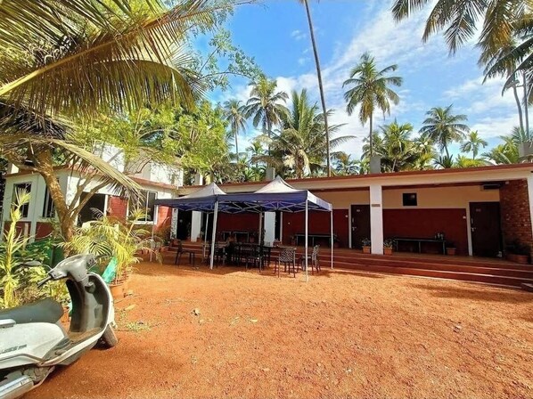 A covered outdoor area with tables and chairs, palm trees, and a dirt ground. - Soham Beach Resort, Diveagar (Shrivardhan)