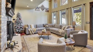Ein geräumiges Wohnzimmer mit einer großen, grauen Eckcouch, einem kleinen runden Couchtisch und einem Kamin. In der Ecke steht ein festlich geschmückter Weihnachtsbaum.