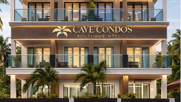 Un hotel de varios pisos con balcones, un cartel que dice 'CAYE CONDOS BOUTIQUE HOTEL' y palmeras en primer plano.