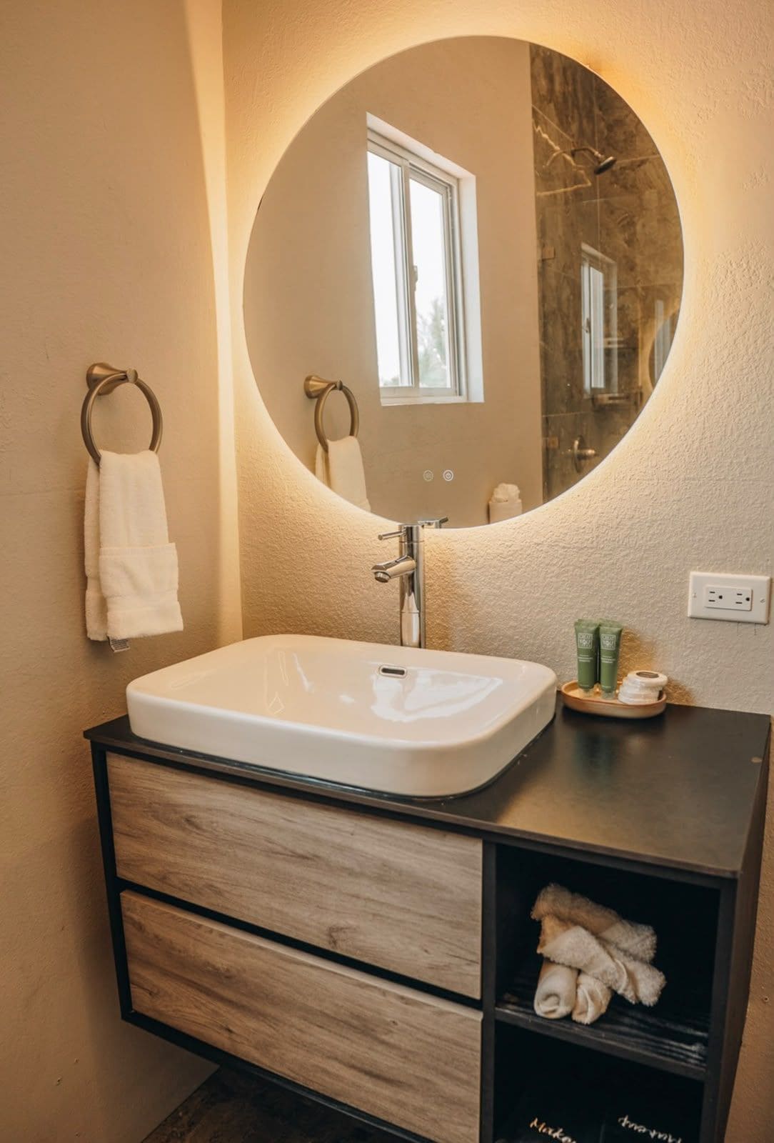 Un baño con un espejo ovalado grande, un lavamanos blanco y un tocador de madera con cajones.