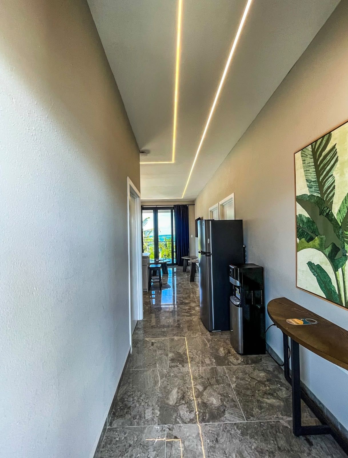 A corridor with a kitchen area, including a refrigerator, coffee machine, and a dining table with chairs.