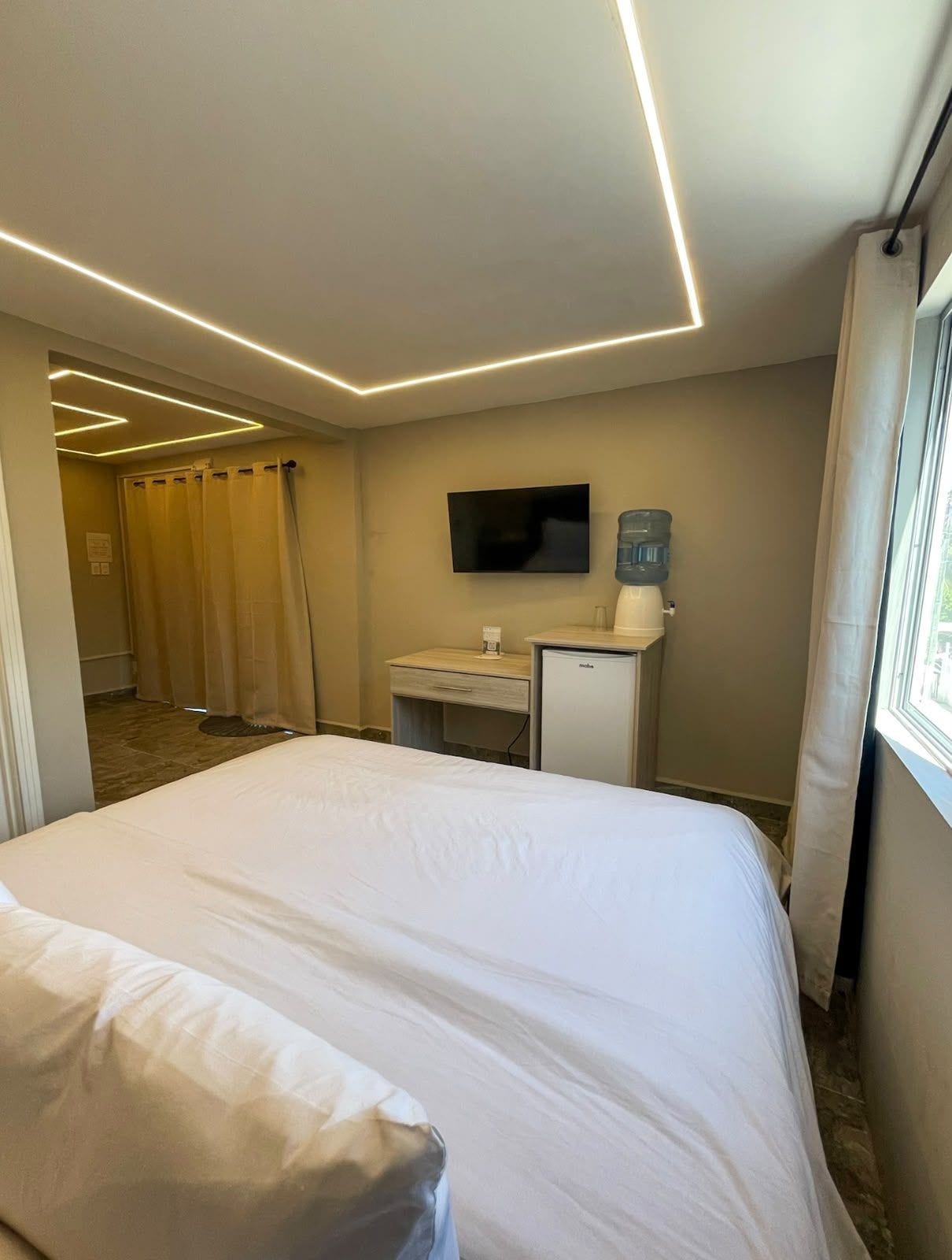 A hotel room with a bed, a desk with a water dispenser, a TV, and a shower area.