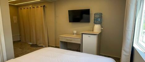 A hotel room with a bed, a desk with a water dispenser, a TV, and a shower area.