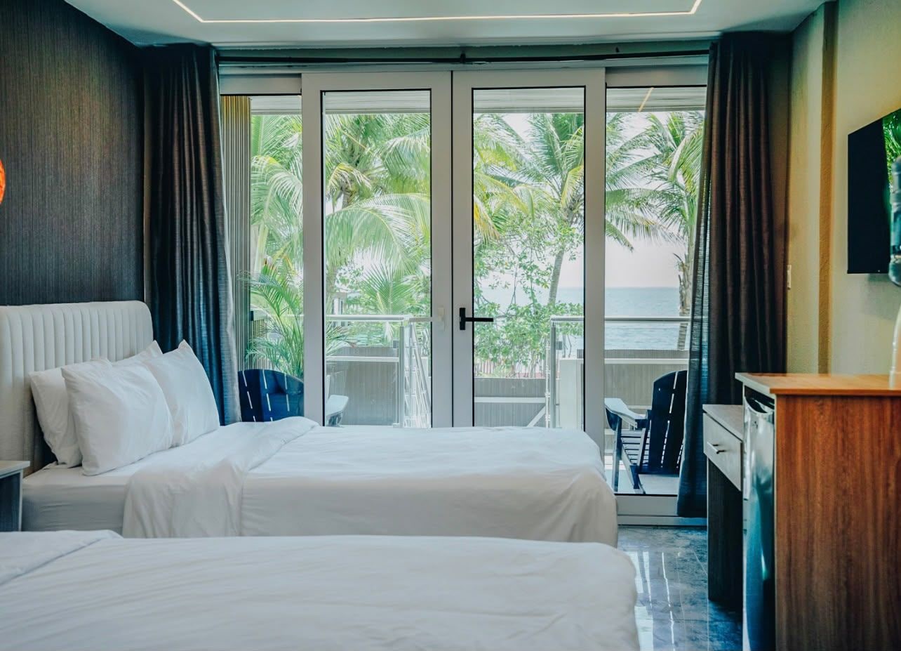 Ein Hotelzimmer mit zwei Betten, einem Schreibtisch und Blick auf Palmen durch die Glastüren.