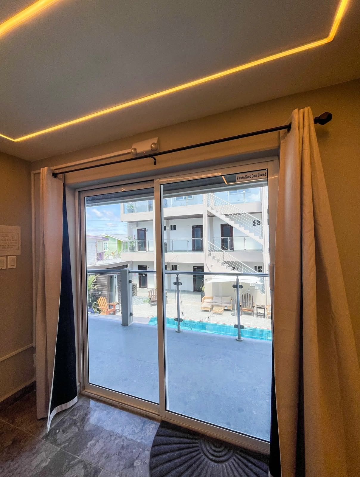A hotel room with a large window, a view of a building with a pool, and a sign indicating the door is closed.