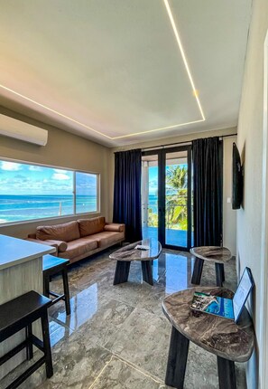 A modern living room with a sofa, a coffee table, and a TV. There is a large window with a view of the ocean. - Caye Caulker Condos By Barefoot (Caye Caulker)