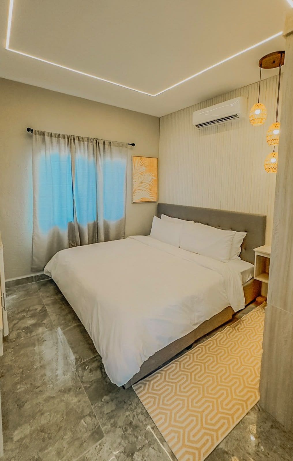 A modern bedroom with a large bed, a bedside table, an air conditioning unit, and a patterned rug.