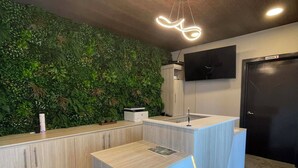 Un mostrador de recepción con detalles de madera, una pared verde con plantas, un televisor de pantalla plana montado y una puerta negra.