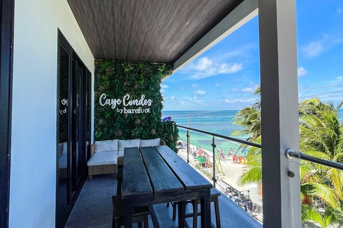 Un espacio con mesas y sillas al aire libre con vista al océano, una pared con vegetación y un letrero que dice 'Caye Condos by barefoot'.