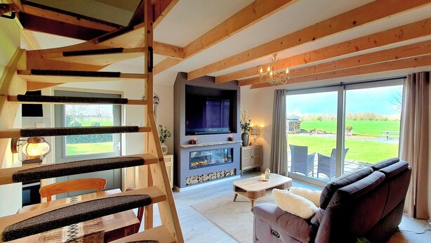 A cozy living room with a fireplace, a television, and a large window offering a view of the outdoors. - Cottage the Bird's Nest with jacuzzi (Oldekerk)