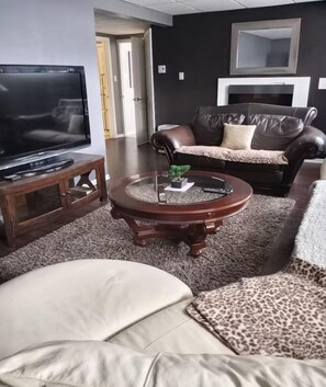 Una sala de estar con una televisión sobre un mueble bajo, una mesa de centro redonda con una planta, un sofá de cuero y un sillón blanco.