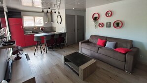 A modern kitchen with red cabinets, a dining area with stools, a brown sofa with red and gray cushions, and a small wooden coffee table. - Gîte 2 **, 55m², 2/4 persons, quiet, 20 min from the beaches Pornic, st Brévin (Vue)