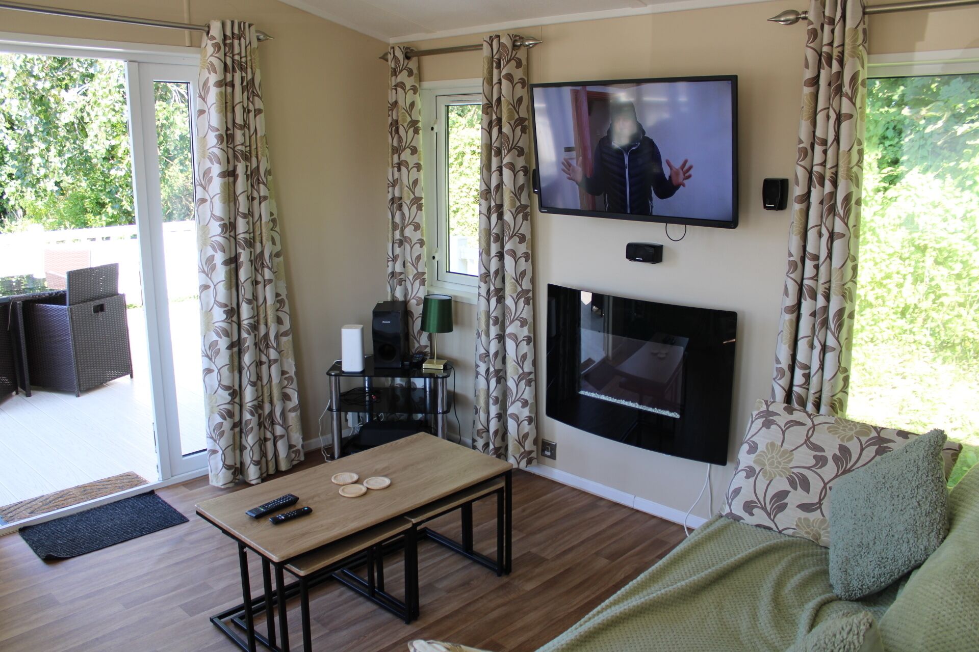 A living room with a fireplace, a television mounted on the wall, a dining table with remote controls, and a sofa with a green throw.