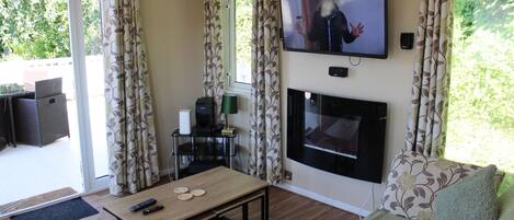 A living room with a fireplace, a television mounted on the wall, a dining table with remote controls, and a sofa with a green throw.