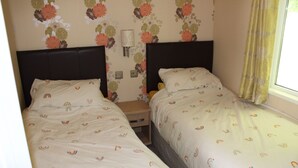Una habitación con dos camas, cada una con una colcha de diseño arcoíris, una mesita de noche, una lámpara de pared y papel tapiz con motivos florales.