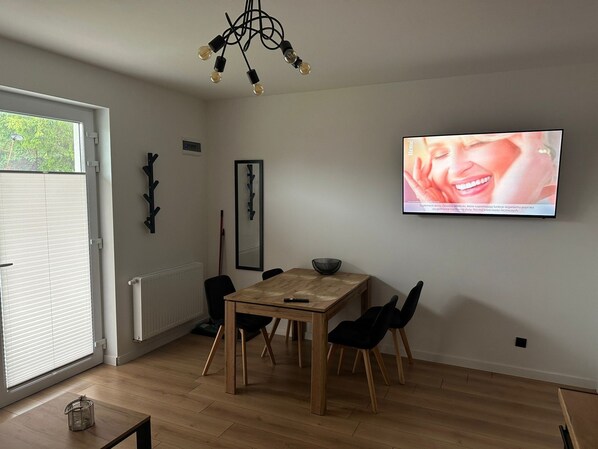 Ein zeitgemäßer Essbereich mit Holztisch und schwarzen Stühlen, einem an der Wand montierten Fernseher und einem Fenster mit Jalousien.