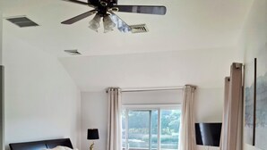 Een slaapkamer met een bed, een bank, een plafondventilator en een groot raam met gordijnen.