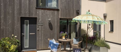 Un edificio moderno con facciata in legno scuro, una porta in vetro e un piccolo spazio esterno con tavolino e sedie in vimini, più un ombrellone decorativo.
