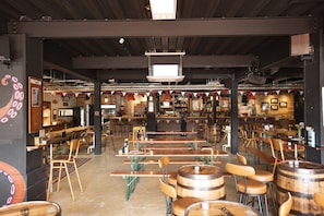 A restaurant interior with wooden tables and chairs, a bar area, and a high ceiling with exposed beams.