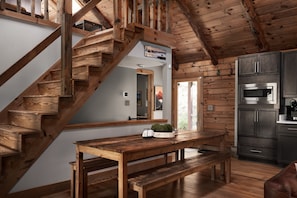 A wooden staircase leading to a loft bedroom, a wooden dining table with benches, and a kitchen area with dark cabinets and a stainless steel oven.