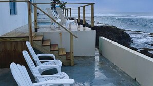 Ein Küstenanwesen mit einer Holztreppe, die zu einem Deck mit weißen Kunststoffstühlen führt.