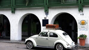 A vintage beige car parked in front of a building with green balconies and arched doorways.