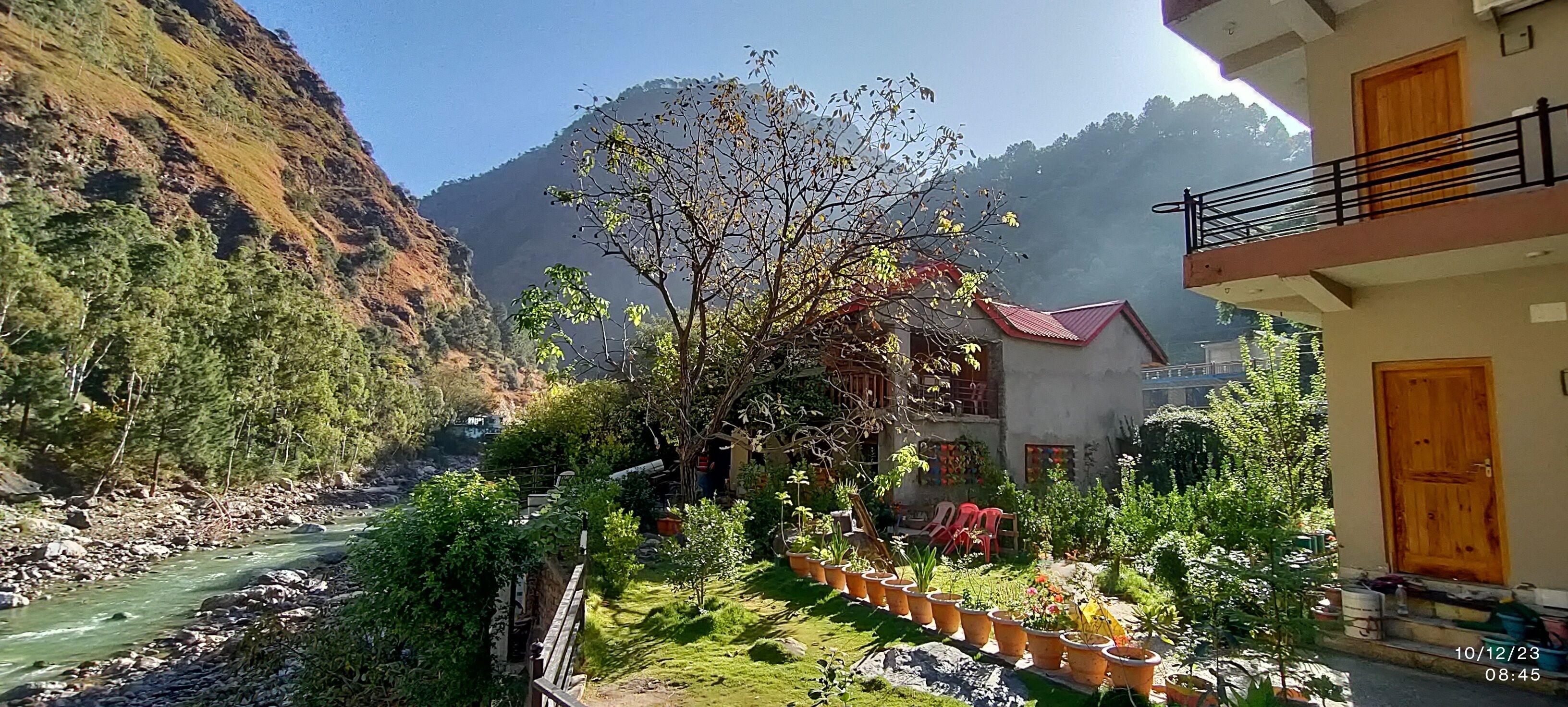 A riverside view with a mountain backdrop, a garden with potted plants, and a building with a red roof.