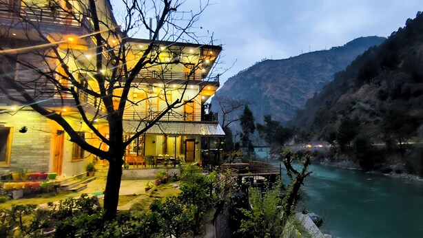 A multi-story building with a balcony overlooking a river, surrounded by greenery and mountains. - Tirthan Grand Riverside  (Sainj)