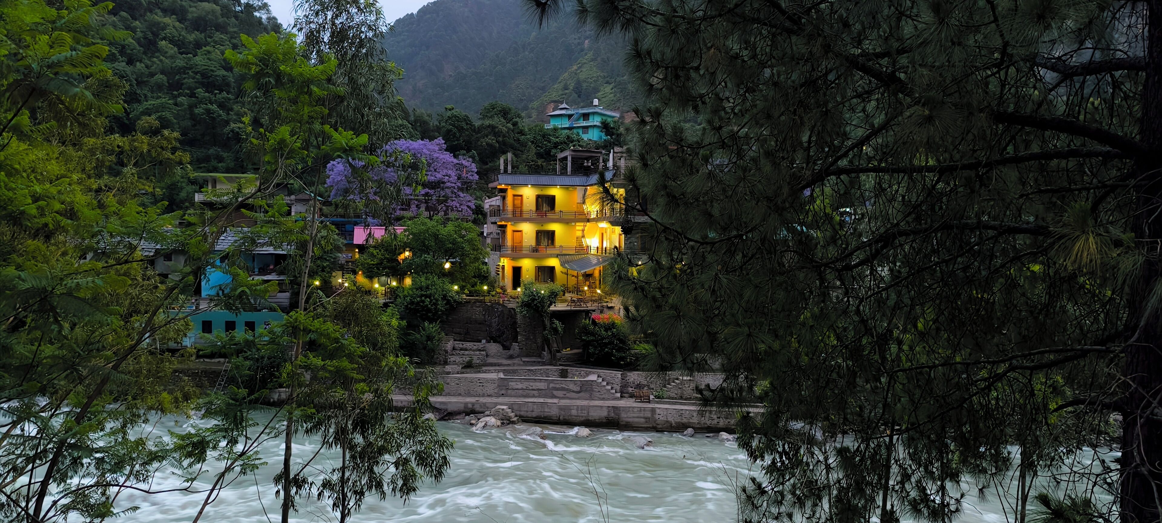 A multi-story building with balconies overlooking a river, surrounded by lush greenery and a mountainous backdrop.