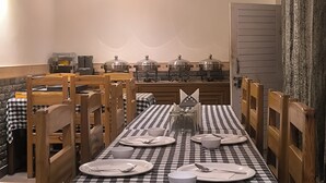 A dining area with a checkered tablecloth, wooden chairs, and a buffet setup with covered dishes. - Tirthan Grand Riverside  (Sainj)