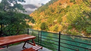 A wooden picnic table with a metal railing overlooking a river and forested hills. - Tirthan Grand Riverside  (Sainj)