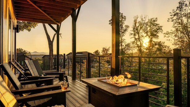 A wooden deck with lounge chairs and a fire feature, overlooking a scenic landscape with trees and distant mountains.