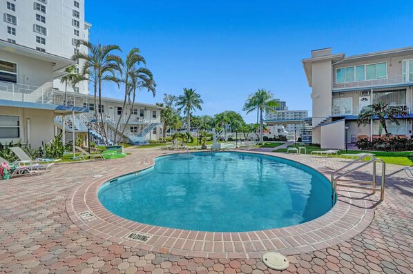A swimming pool with a brick border, surrounded by palm trees and a multi-story building with balconies. - Oceanfront Oasis Charming 1BR Getaway (Fort Lauderdale)